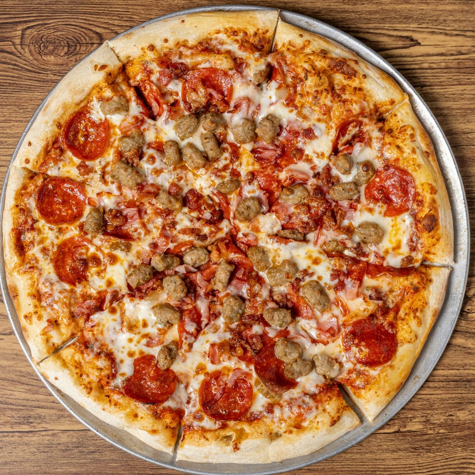 A sliced CD Top Shelf pizza with pepperoni, sausage, and bacon on a wooden table.